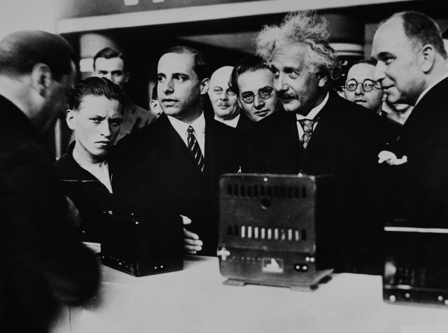 Einstein stands among a crowd at a table with equipment on it.